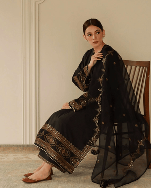 Woman in black embroidered ethnic dress with gold detailing, sitting on a wooden chair indoors.