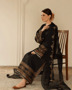 Woman in black embroidered Pakistani formal dress with gold details, sitting on chair