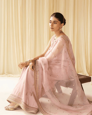 Woman in pastel pink embroidered saree with gold jewelry, sitting indoors on a bench