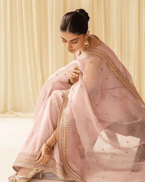 Woman in pastel pink embroidered saree, gold heels, and traditional jewelry, sitting indoors