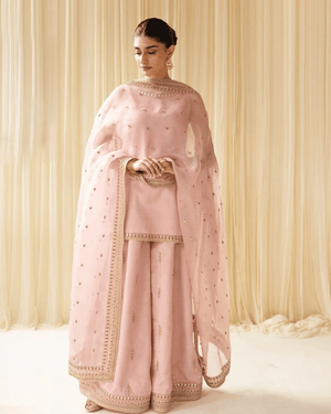 Woman in pastel pink embroidered sharara suit with dupatta, standing by cream curtains