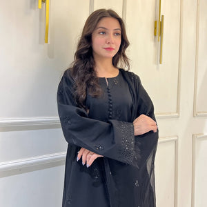 Woman in black embroidered abaya dress with wavy hair, standing against white panel wall
