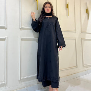 Woman in black embroidered long kurta with matching pants, standing indoors by white wall