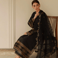 Woman in black embroidered ethnic dress with gold detailing, sitting on a wooden chair indoors.