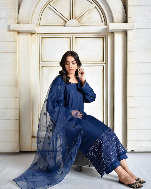 Woman in azure blue embroidered suit and dupatta seated by an arched white door