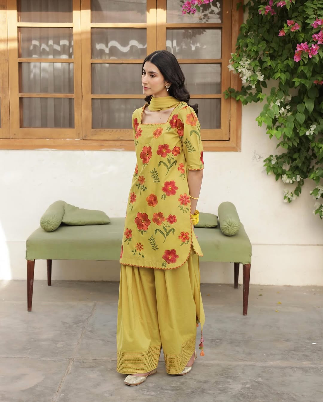 Woman in yellow floral kurta and palazzo pants in courtyard with green bench and flowers