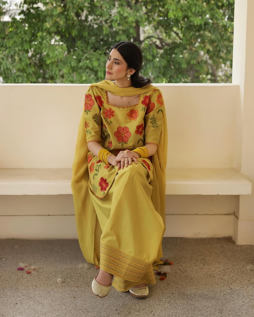 Woman in yellow floral salwar kameez sitting on a bench, trees in background